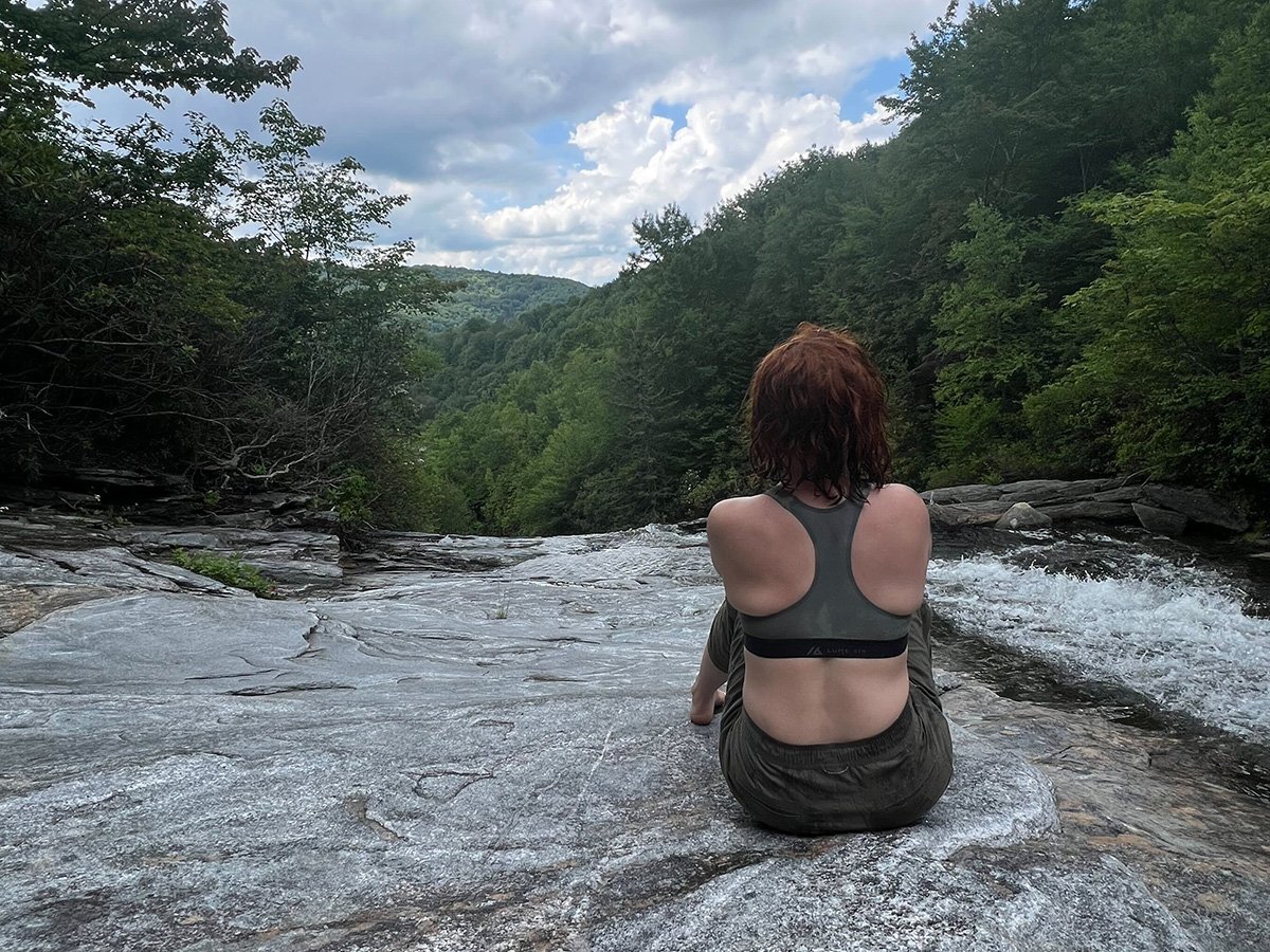 bra_lume2_landscape Woman sitting on rocks next to a river looking over the view. She is wearing a sports bra and pants, and facing away from the camera.