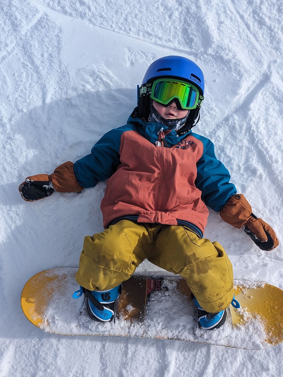 Kid with snowboard on, lying in the snow, wearing a Giro Crue helmet.