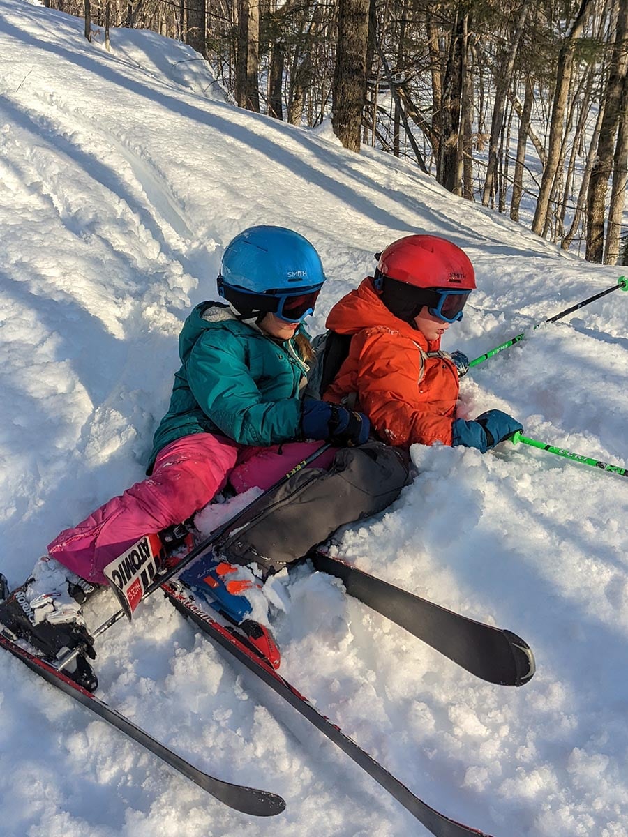Two kids lying in the snow wearing skis, ski clothes, and their Smith Glide Jr. MIPS helmets.
