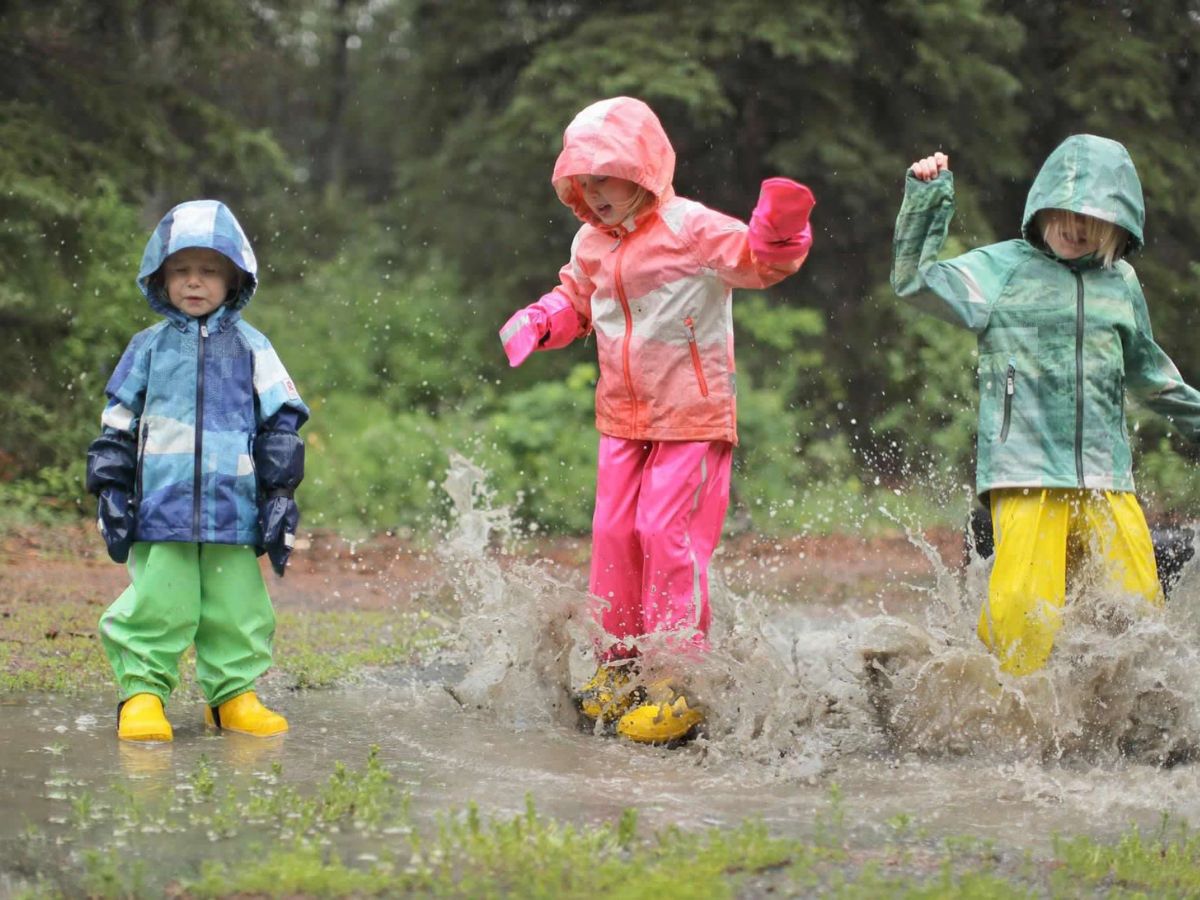 ReimaRainbigpuddlesplash Three kids in bright reima rain gear jump in a puddle.