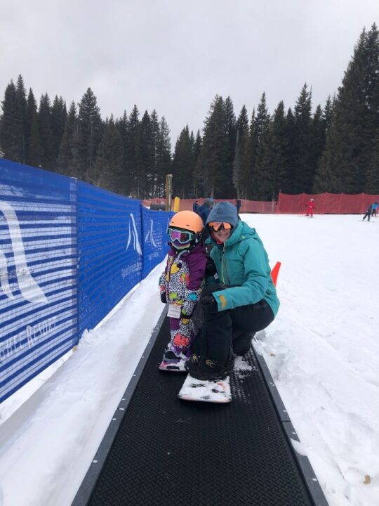 Woman wearing Burton bib snowpants riding magic carpet with toddler.