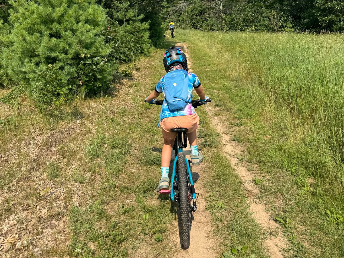 A child rides a mountain bike with a blue Osprey Moki backpack on.