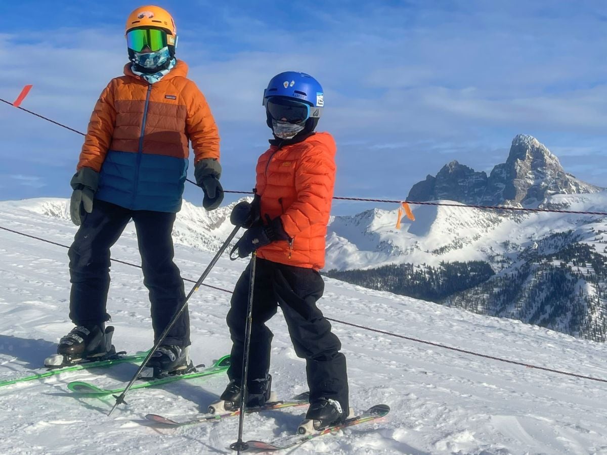 Two boys in helmets stand on skis with a mountain in the background.