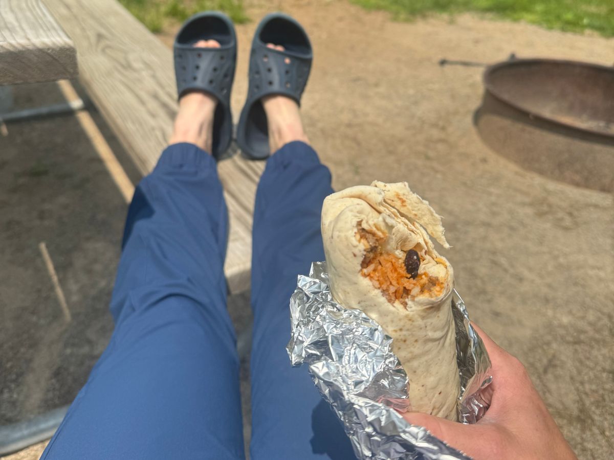 hikingpantst9 A pair of legs wearing blue joggers with feet resting on a picnic table bench and a hand holding a burrito.