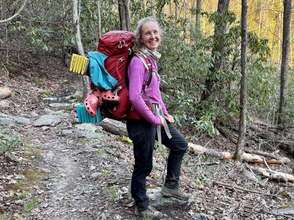 hikingpantst9clamberpant A woman with a pink shirt, black pants and a backpack full of gear smiles for a photo.