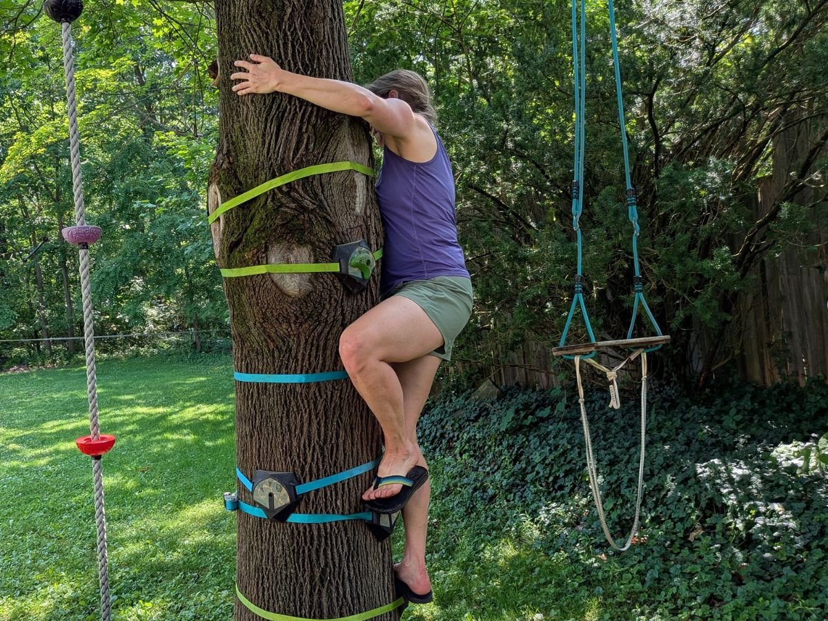 hikingpantst9cordillerashort A woman with a purple tank top, green shorts, and flip flops climbs up holds on a tree.