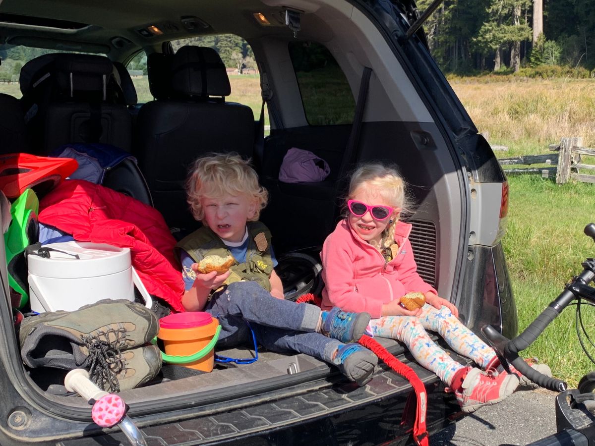 Two kids in the trunk of a van eating muffins. The girl has pink sunglasses and the boy has blue shoes