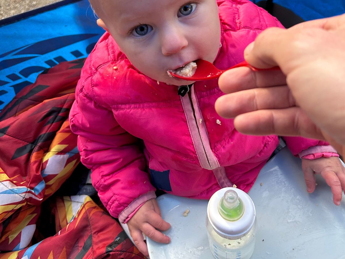 Baby in a pink puffy jacket being fed cereal with a red spoon