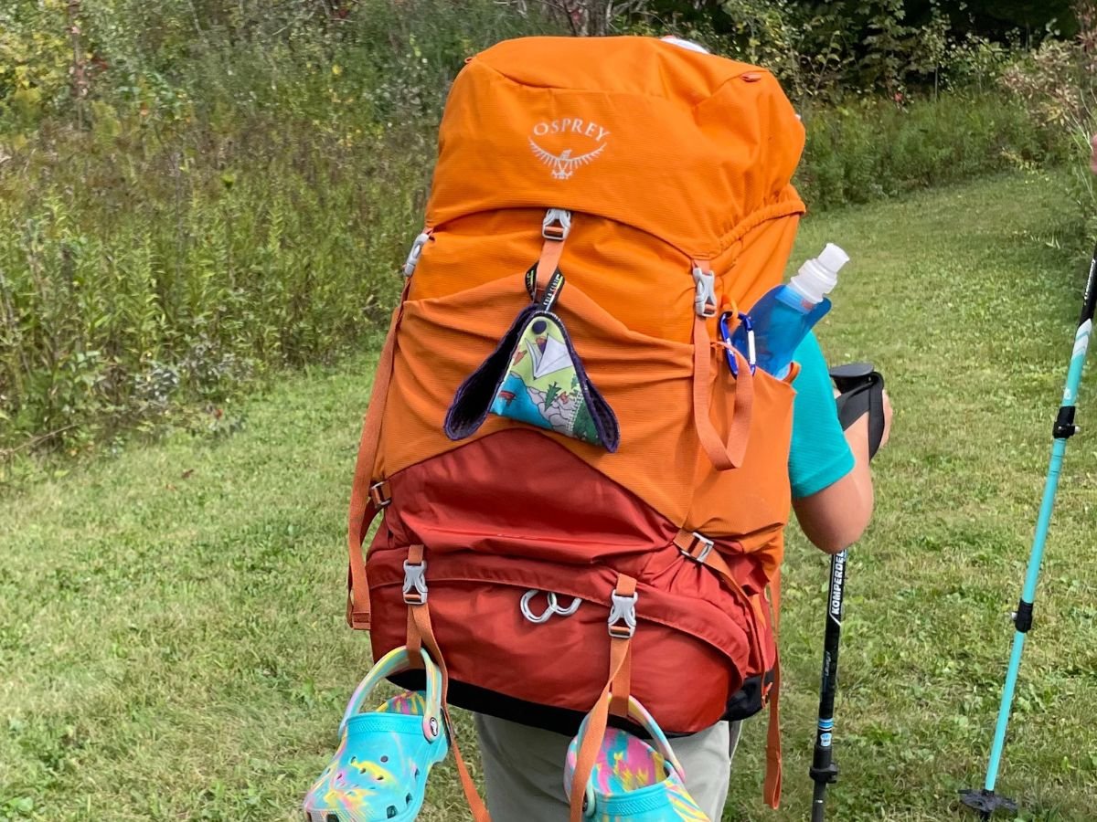 A child is hiking with an orange Osprey pack with a Kula cloth and Crocs hanging off the back.