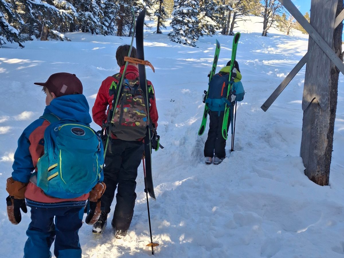 3 kids are hiking up a snowy hill with backpacks and skis on their backs. 