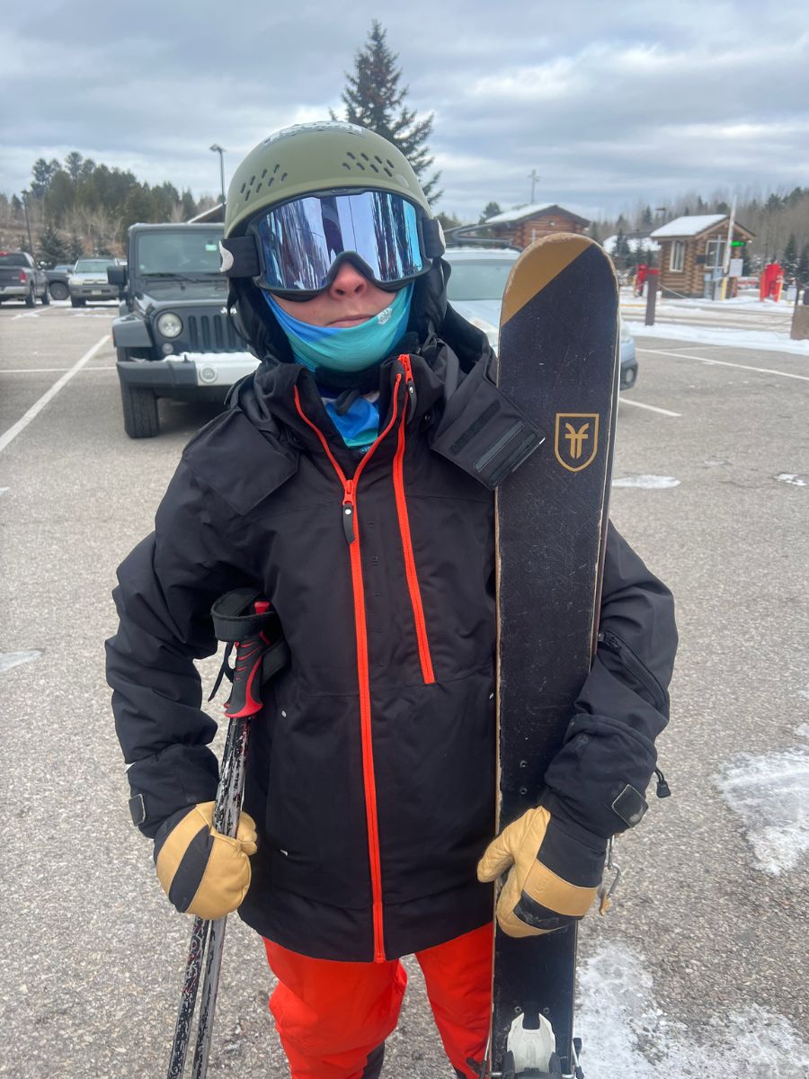 WomenOwnedGear_ChopperMill A kid in helmet and ski goggles holds his skis and poles in a parking lot. He's wearing a beautiful pair of leather winter mittens from Chopper Mill.