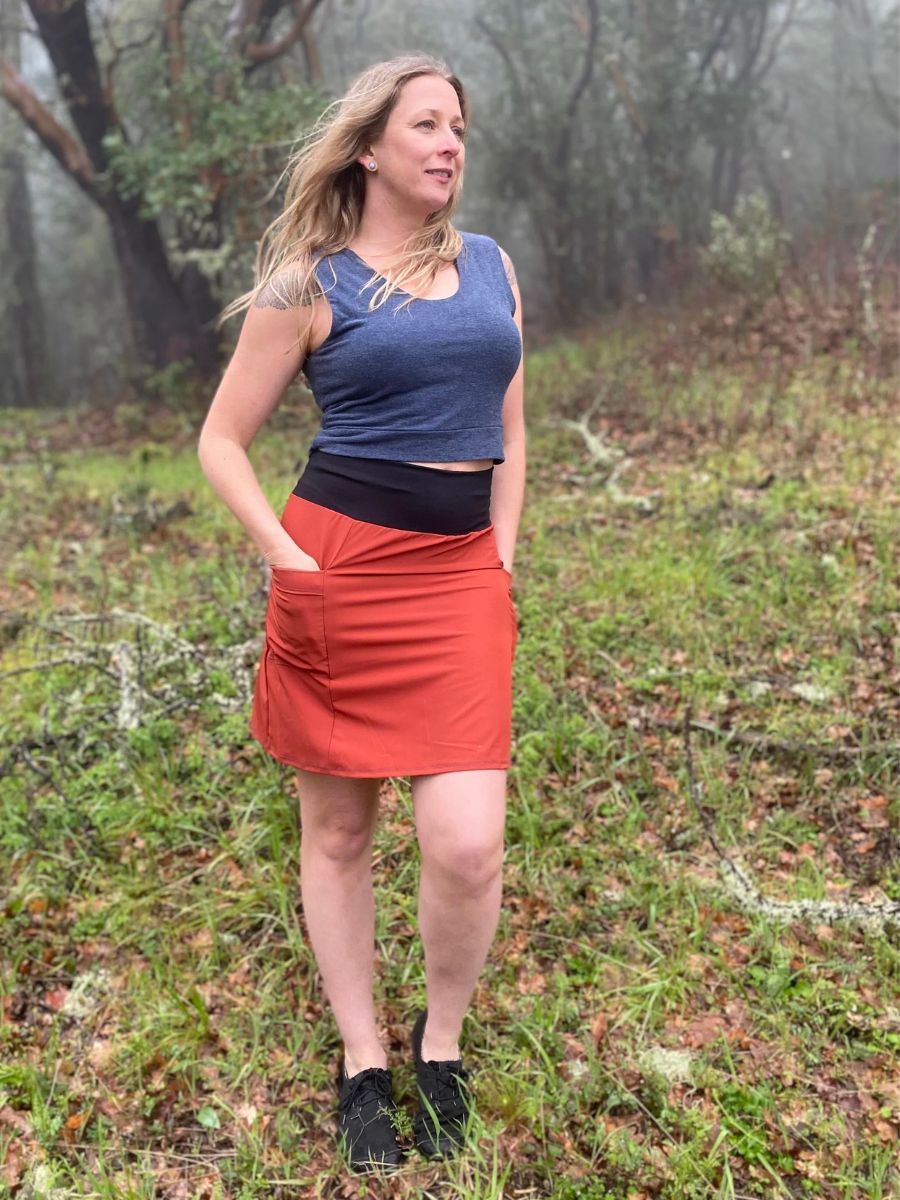 A woman in a grey shirt and orange skirt stands in a foggy forest.