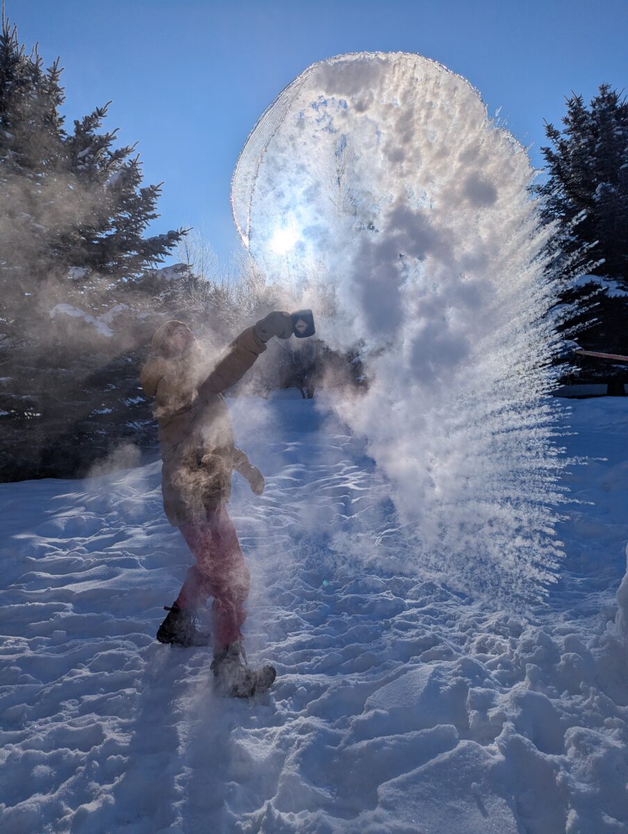 A shower of snow steam backlit by the sun with a boy in the midst.