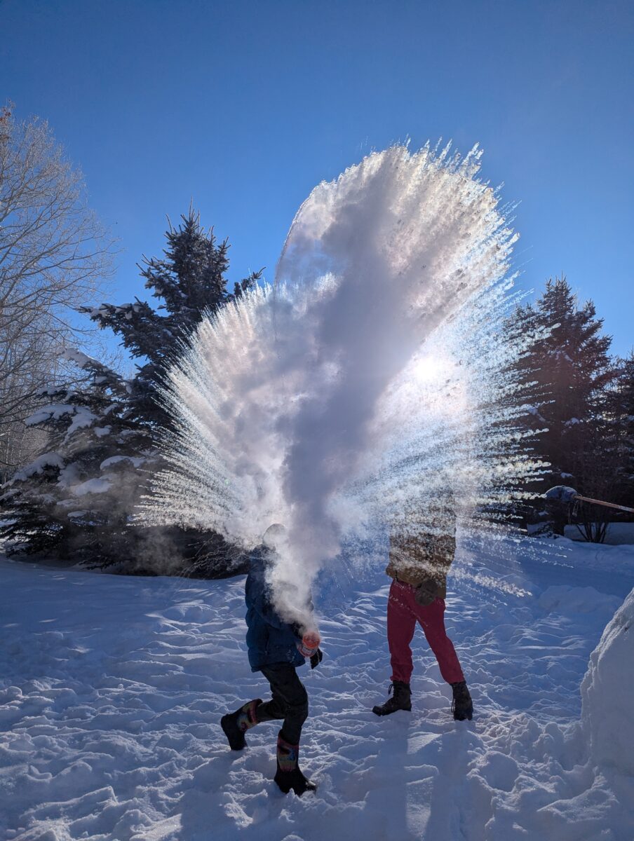 A shower of snow steam backlit by the sun with a boy in the midst.