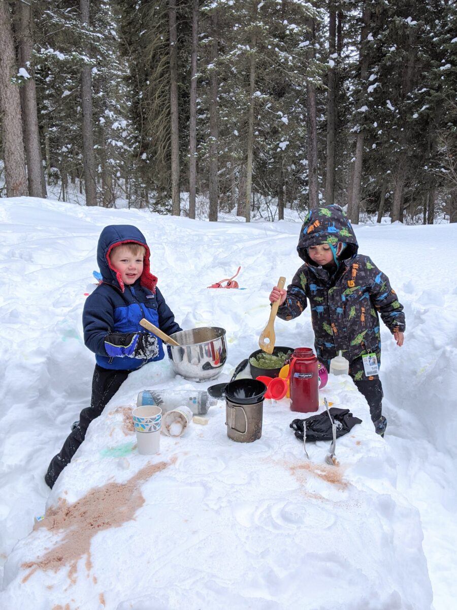 Two children dressed in snow clothes playing with a snow kitchen,