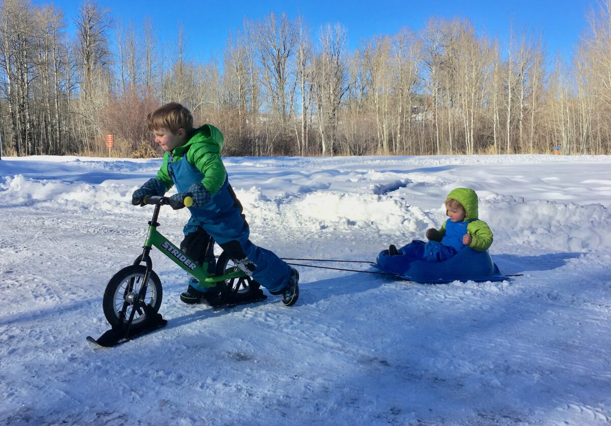 A child on a ski strider bike pulling another child in a sled on the snow.