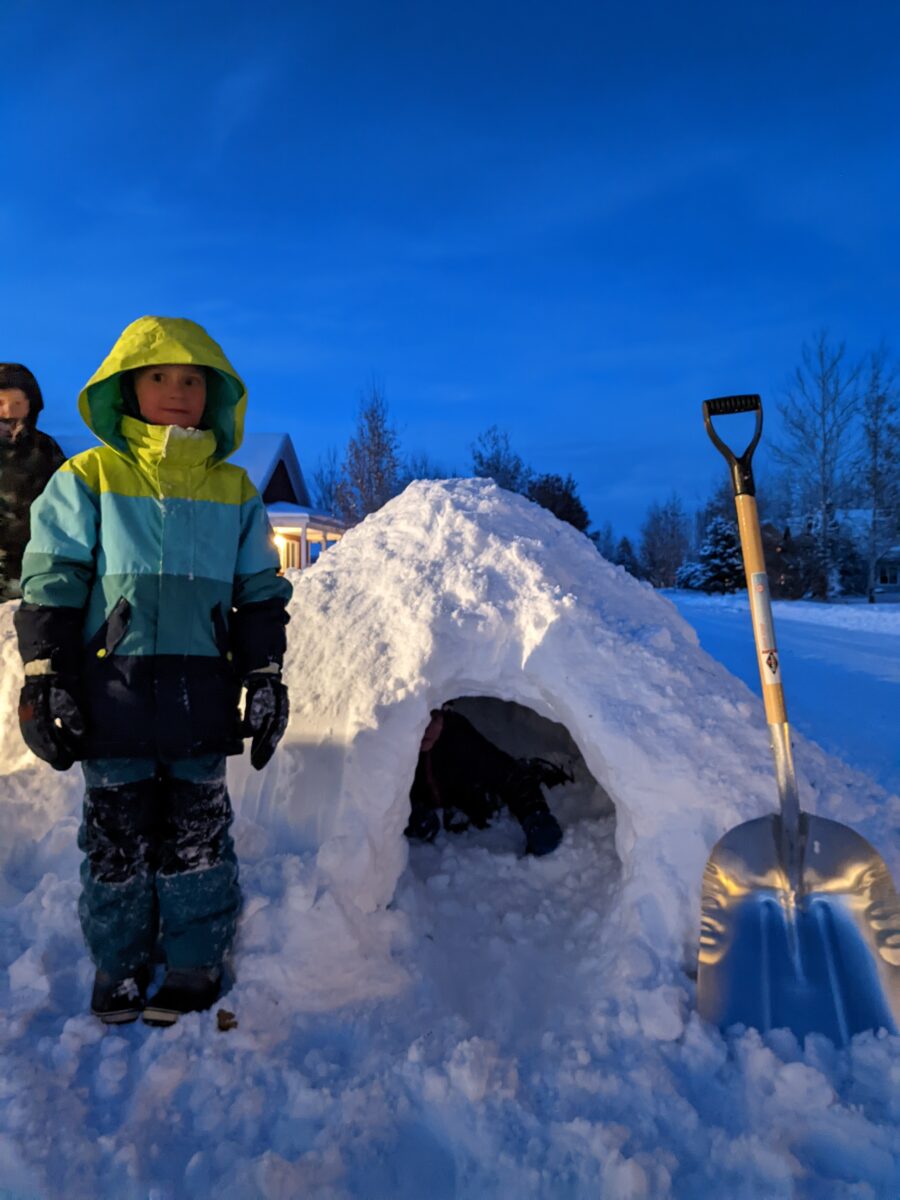 A kid standing next to a snow cave with a shovel.