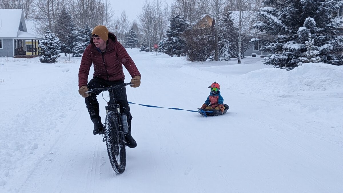 A dad on a bike towing a kid in a sled through a snowy street.