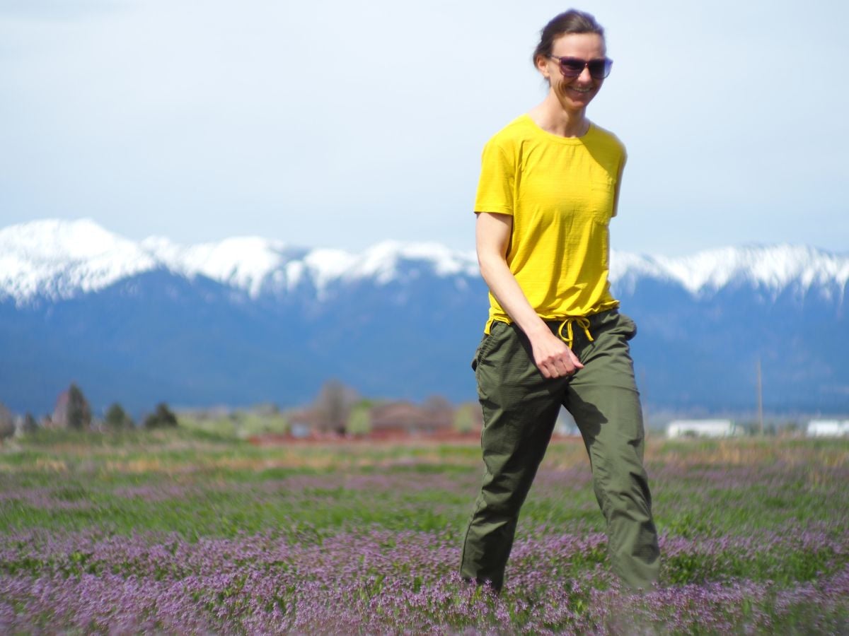 t9sunandswim_1 A woman walks through purple flowers with snow capped mountains in the background with a yellow sun shirt on.