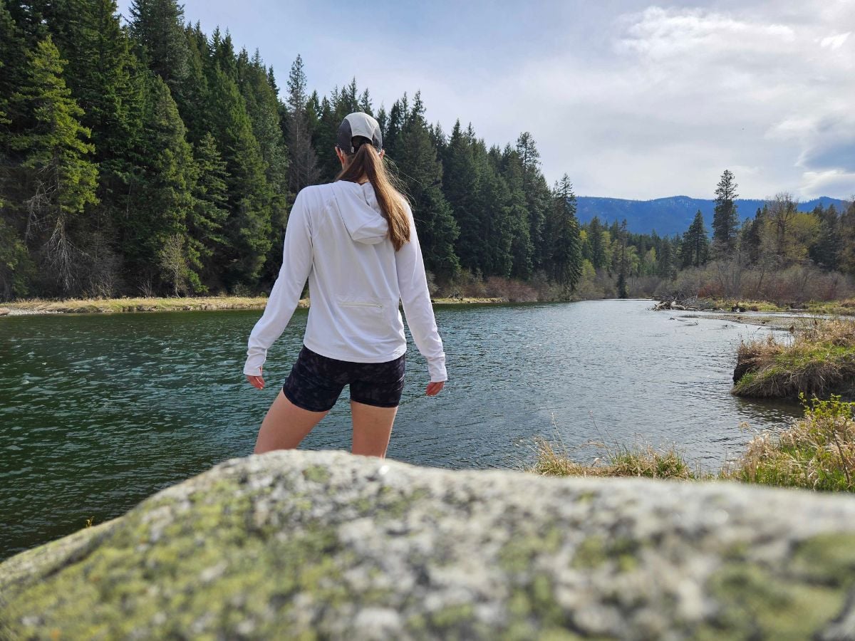t9sunandswim_2 A woman stands in front of a rock by a lake with a white sun hoodie on.