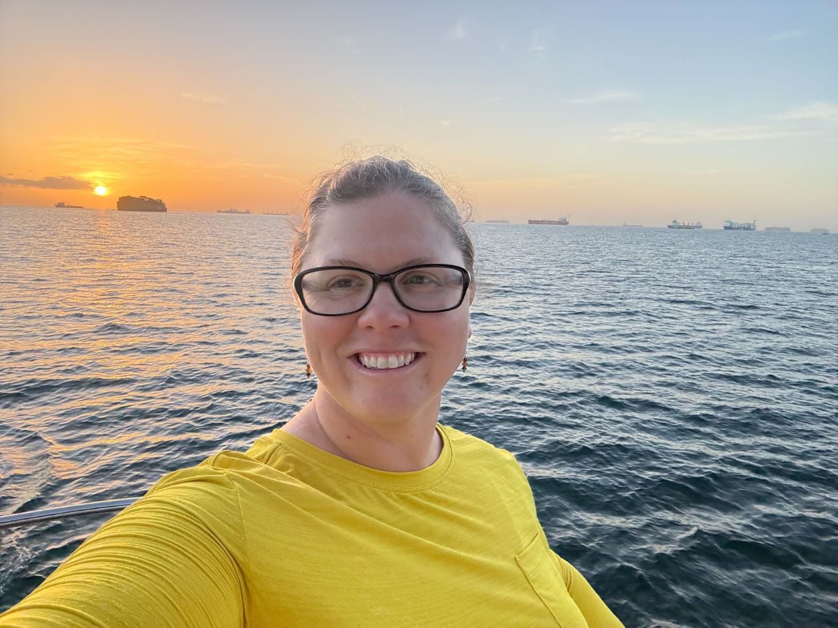 t9sunandswim_7 A woman with glasses and a yellow shirt stands in front of a sunset over the ocean.