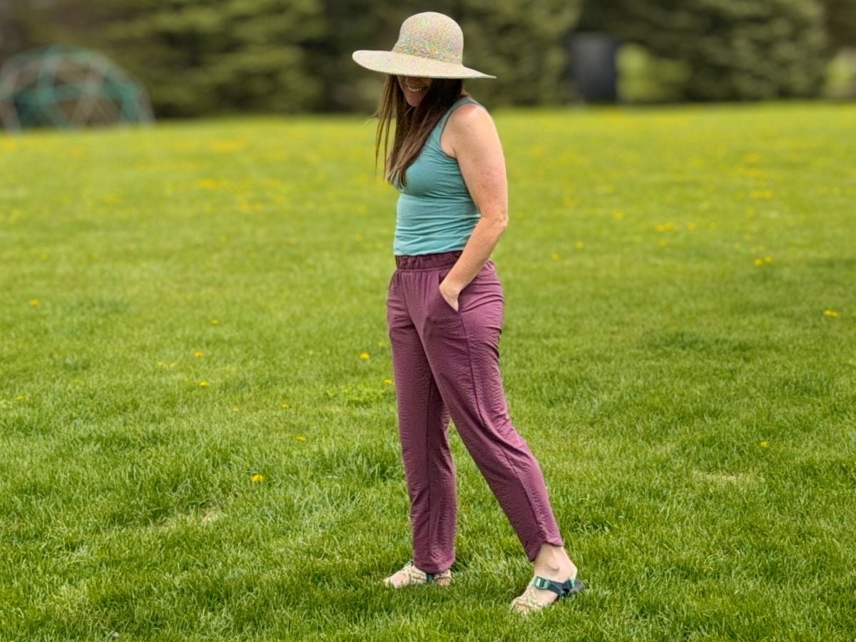 t9sunandswim_8 A woman with a hat, teal tank top and burgundy pants walks in the grass.