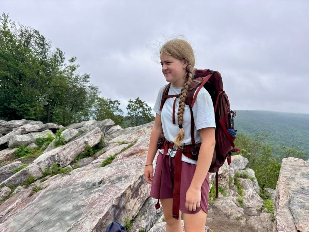 A teenage girl poses in backpacking gear on top of a rocky outcropping.