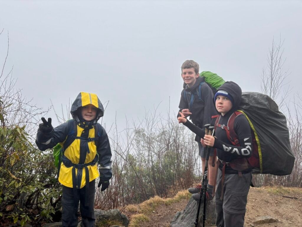Three boys wear backpacking gear, including rain gear, in a foggy mountain setting.