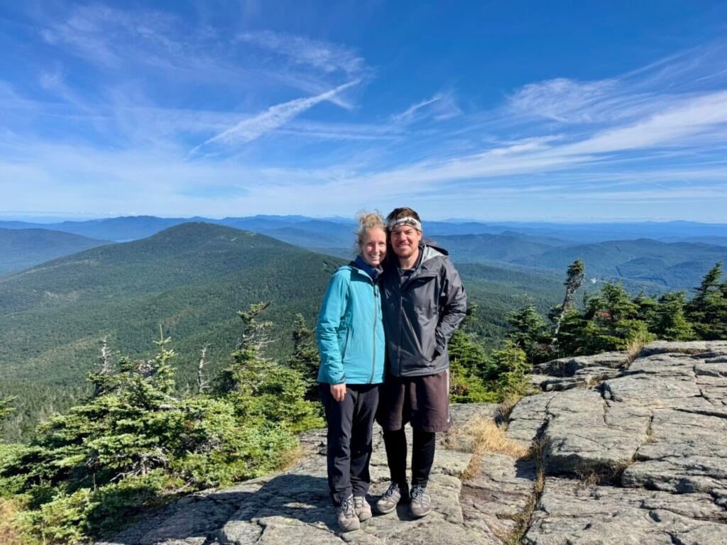 A couple poses in chilly weather gear on top of a mountain beneath blue skies.