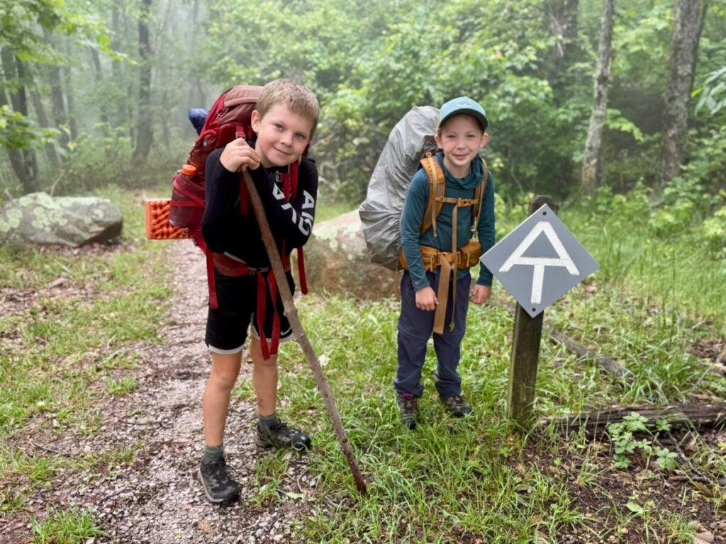 Two kids in backpacking gear pose with an Appalachian Trail sign on a wooded path.