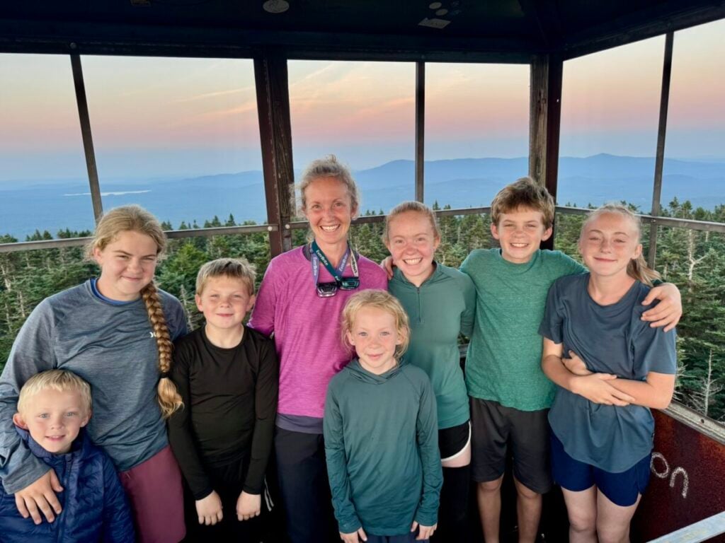 A mother poses with seven children inside a fire tower, with a sunset over mountains visible in the background.