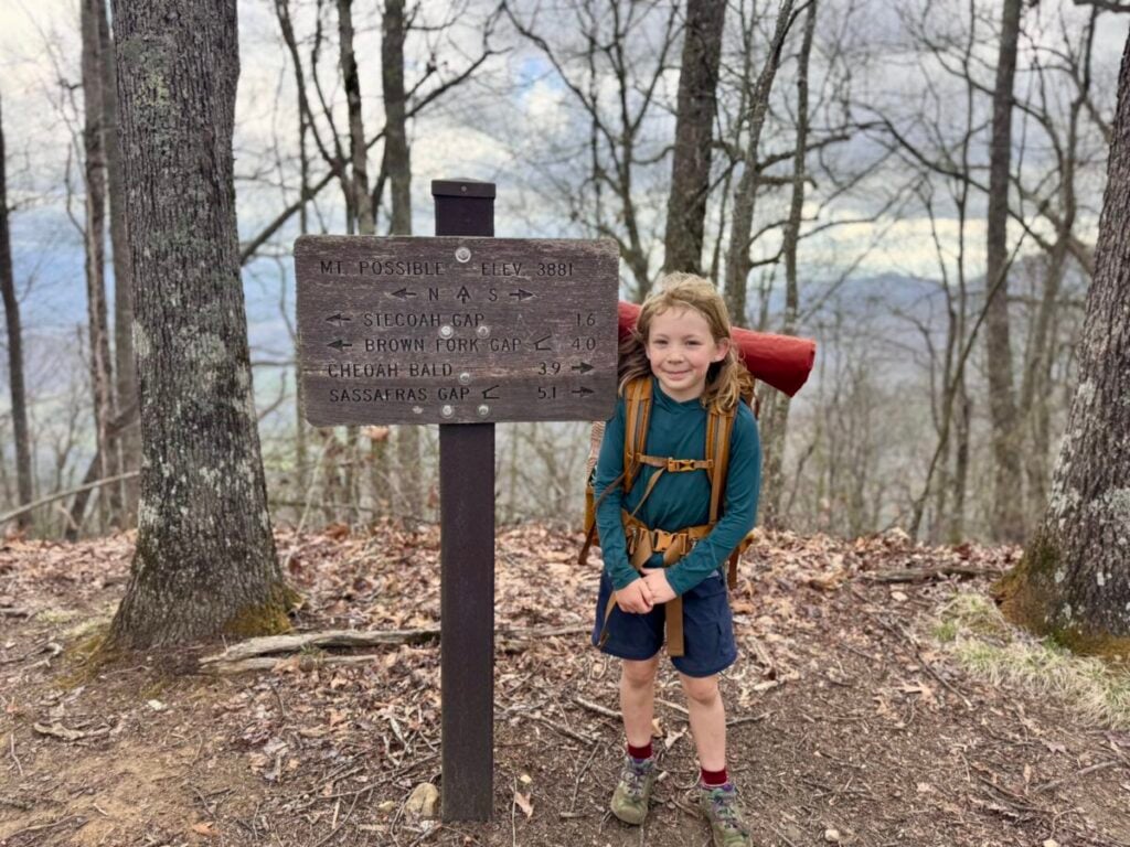 A young girl wearing backpacking gear poses with a sign for Mount Possible.