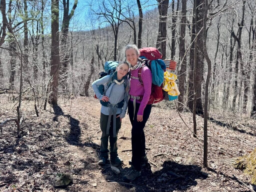 A mother-daughter pair wearing backpacking gear poses on a wooded trail.
