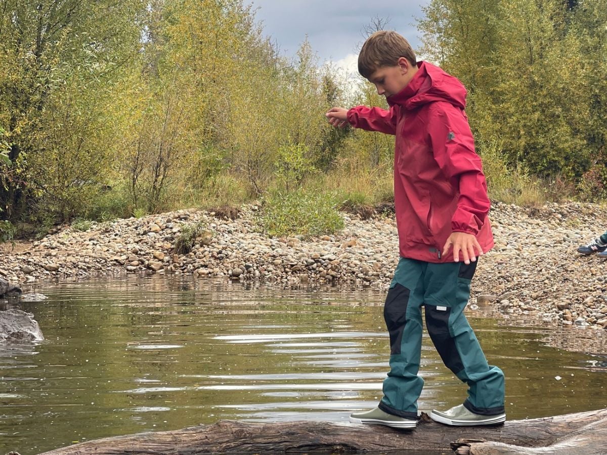ReimaRainSampuPants A young boy with a red jacket and teal pants walks on a log across water