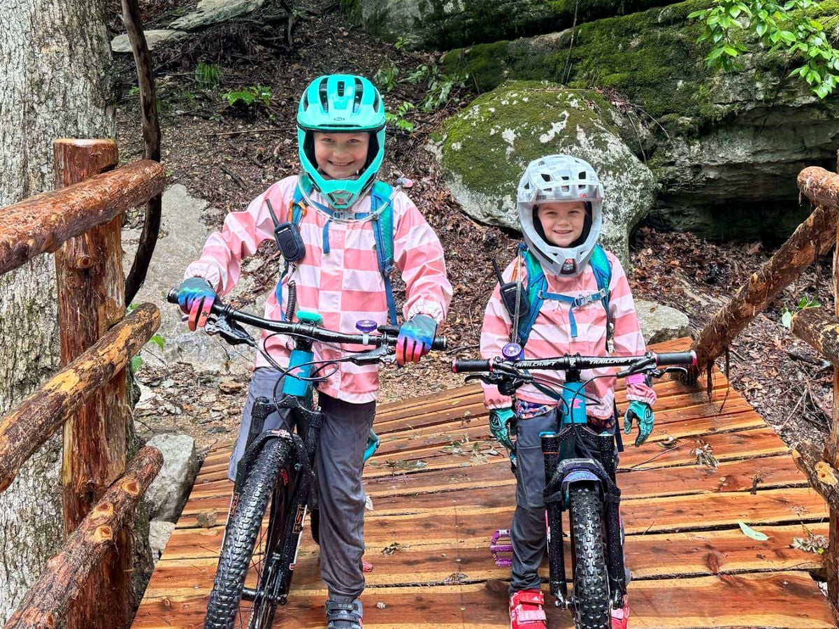 ReimaRainVesiJacketBike Two girls in striped Reima Vesi Rain Jackets wear bike helmets and stand over their bikes.