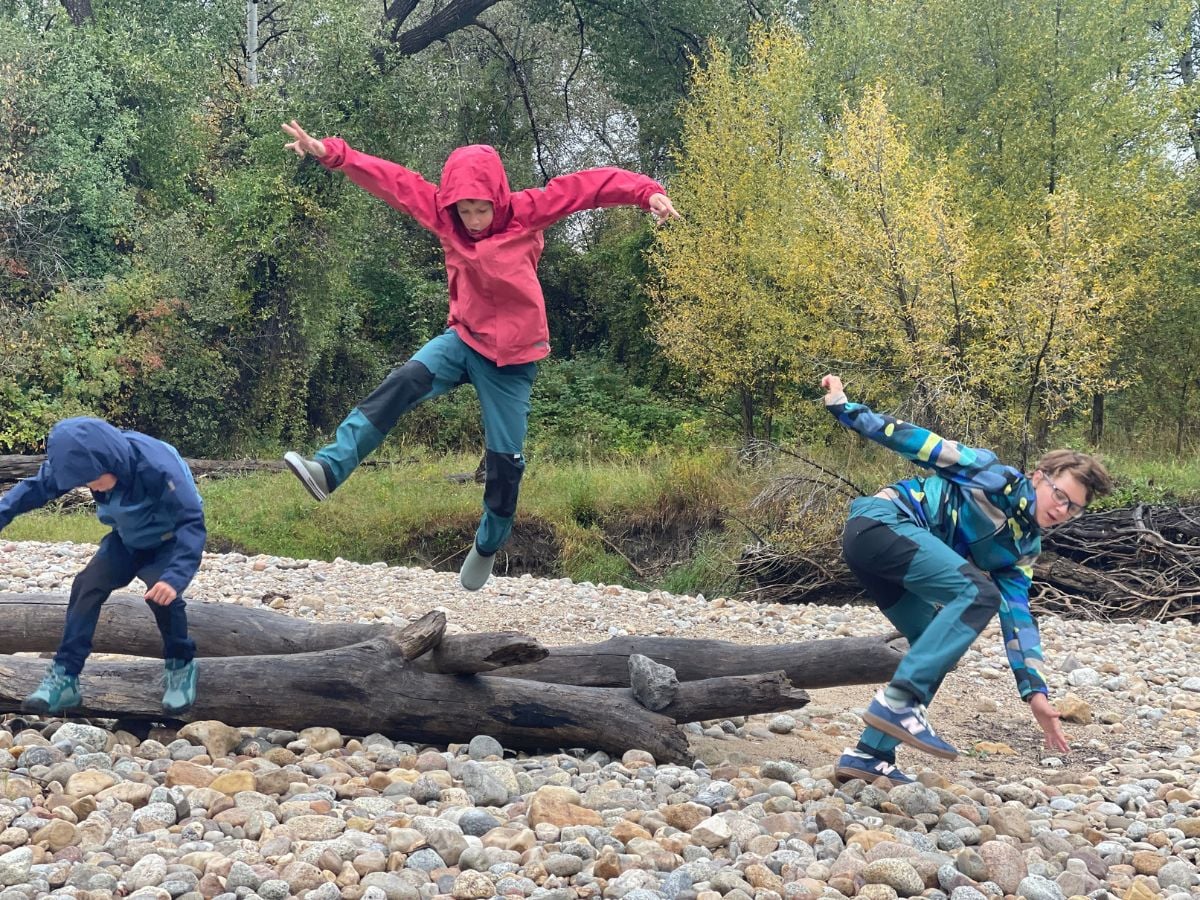 Three boys jump off a log dressed in Reima gear for rain and hiking gear.