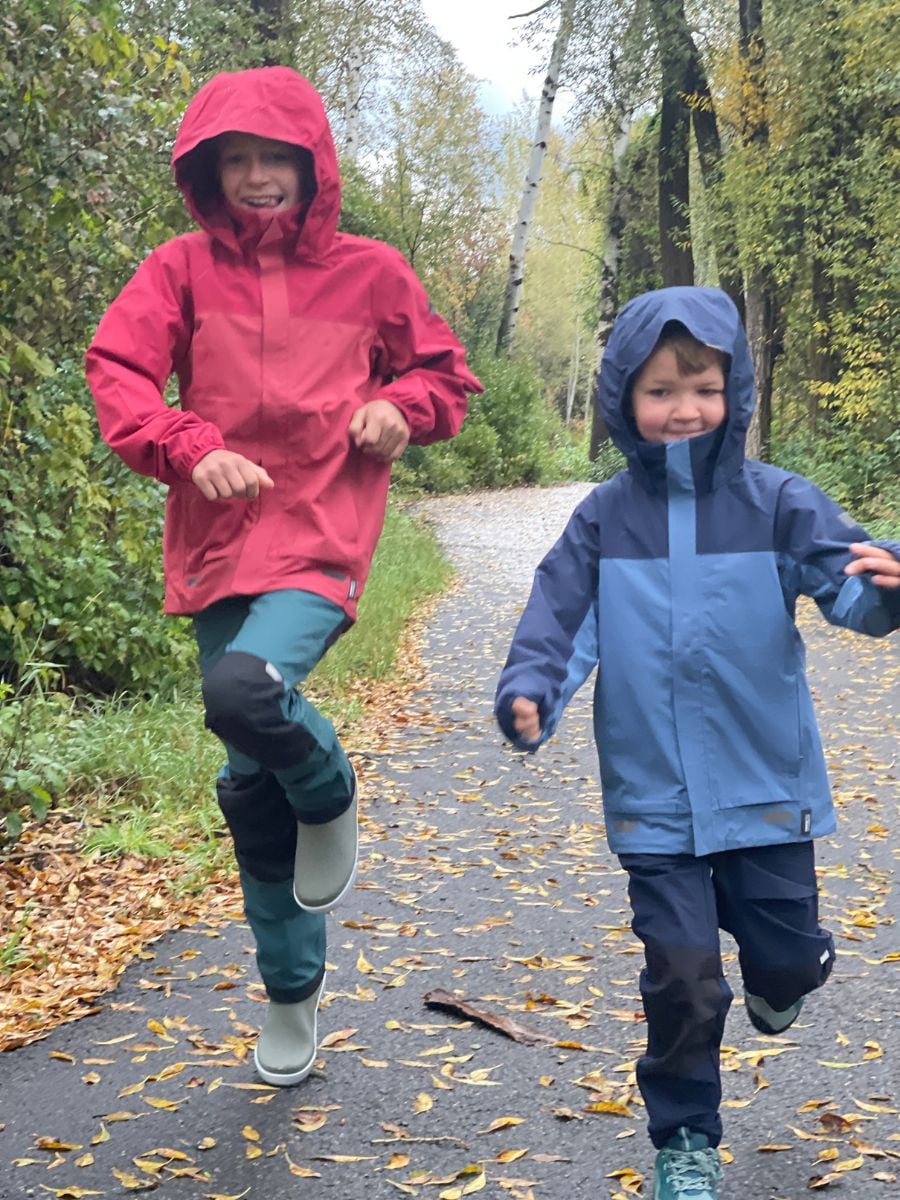 Two boys happily running along a wooded path dressed in rain gear.