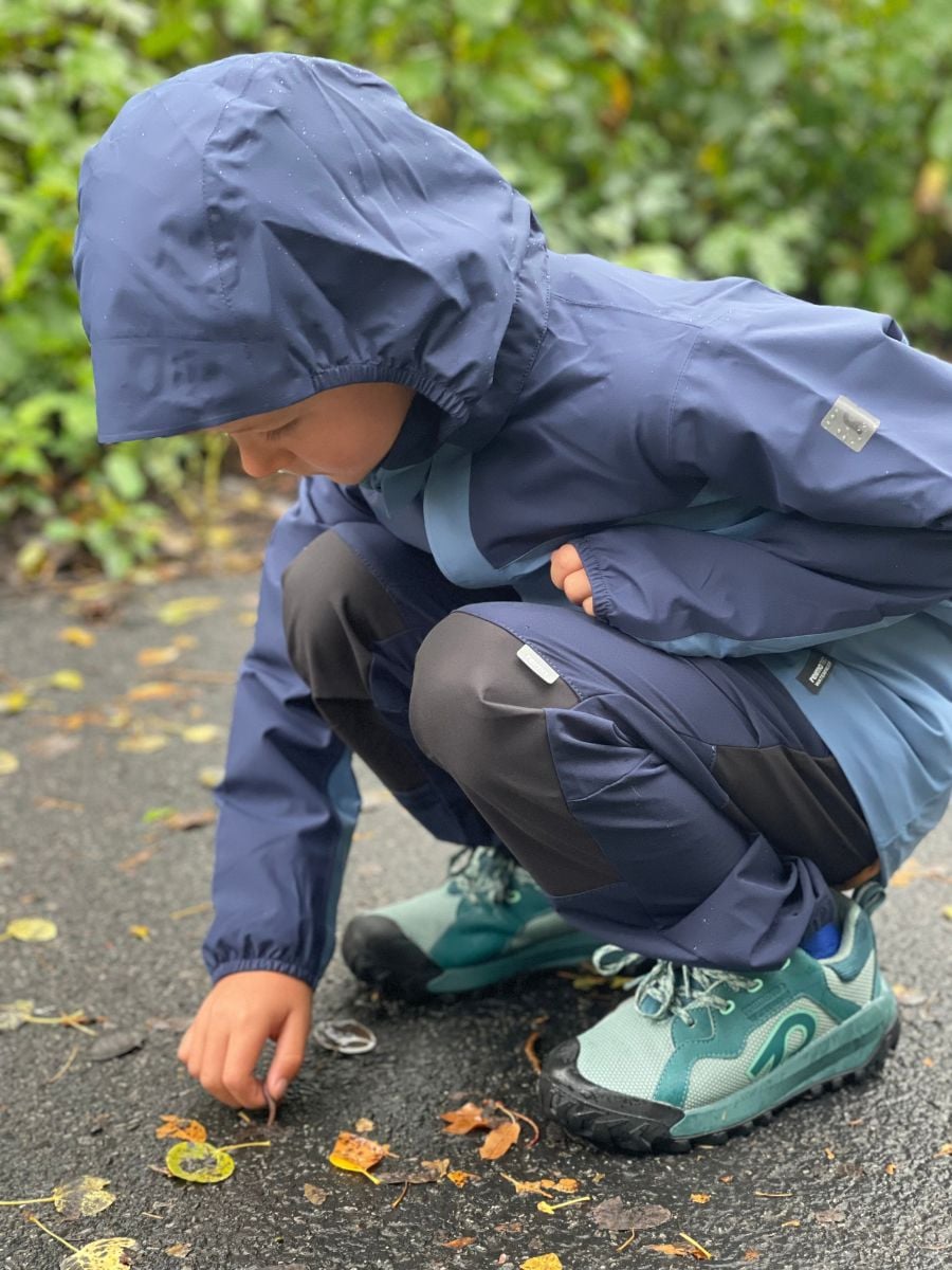 A boy inspects a leaf on a bike path dressed in Reima rain gear and hiking shoes.