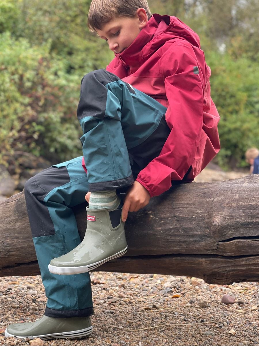 A boy in Reima rain gear puts on a rain boot.