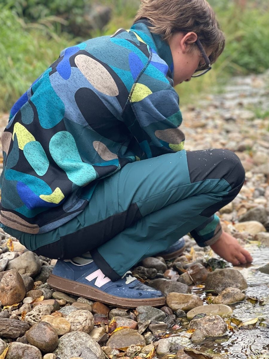 A boy dressed in rain pants and a fleece top inspects a rock in a creek.