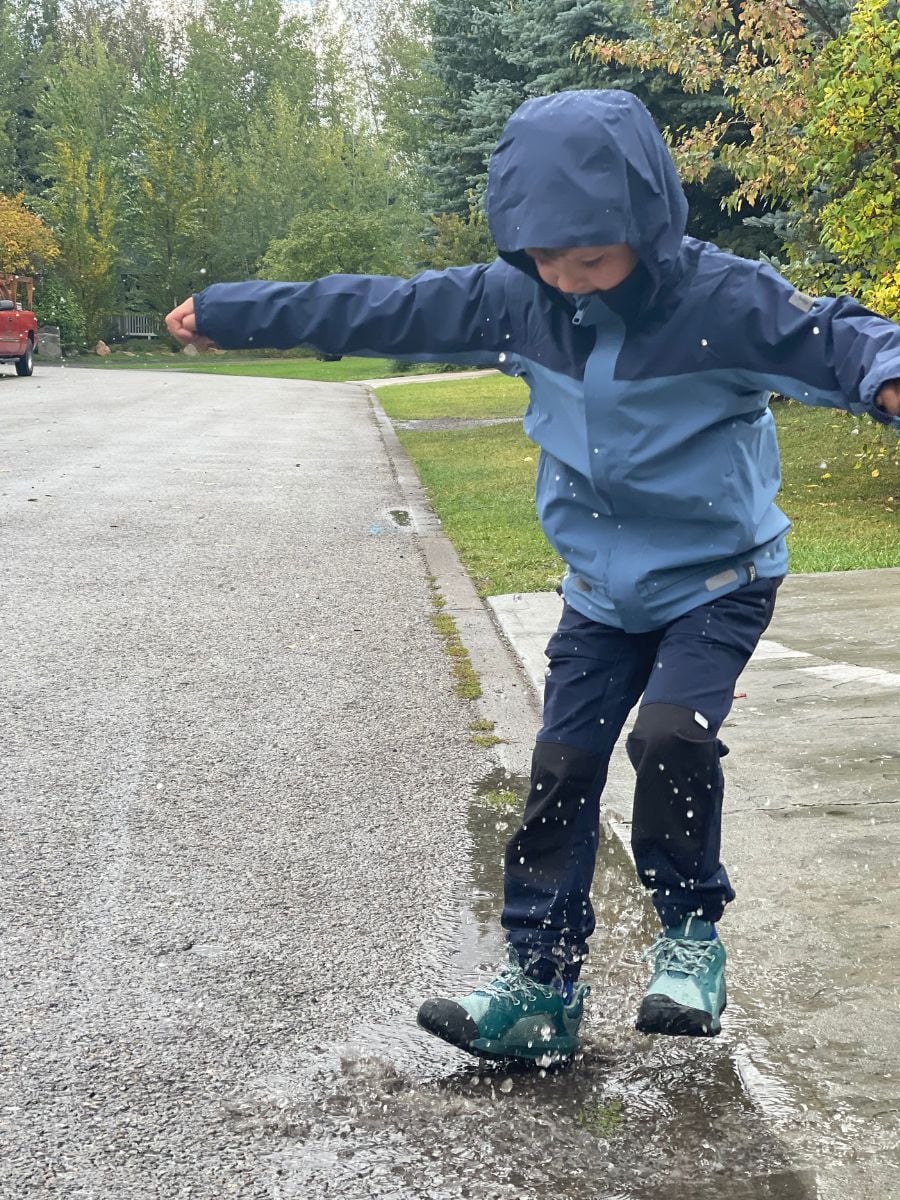 A boy in blue rain gear jumps in a puddle.