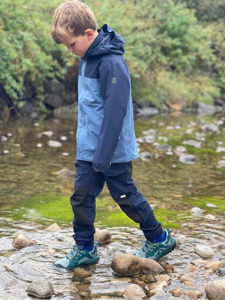 A boy in blue rain gear walks through a creek.