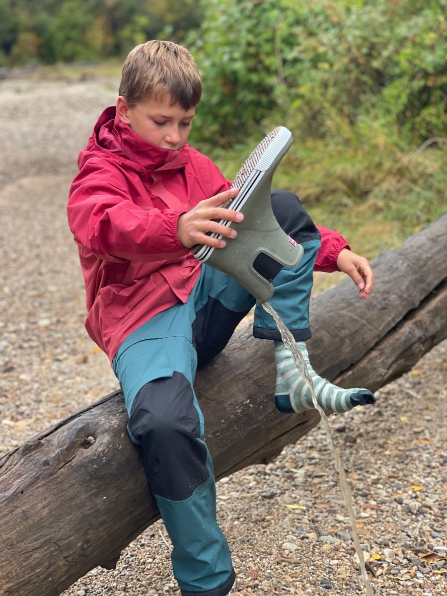 A boy dressed in rain gear dumps water out of his boot.